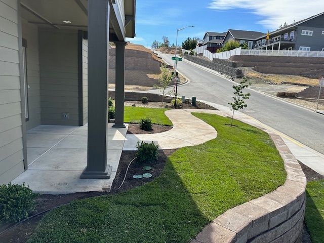 A porch with a walkway leading to a sidewalk in a residential neighborhood with landscaped grass and stone retaining walls.
