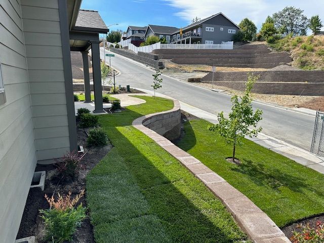 A suburban house with a landscaped lawn, curved stone retaining wall, and a view of a street with neighboring homes.