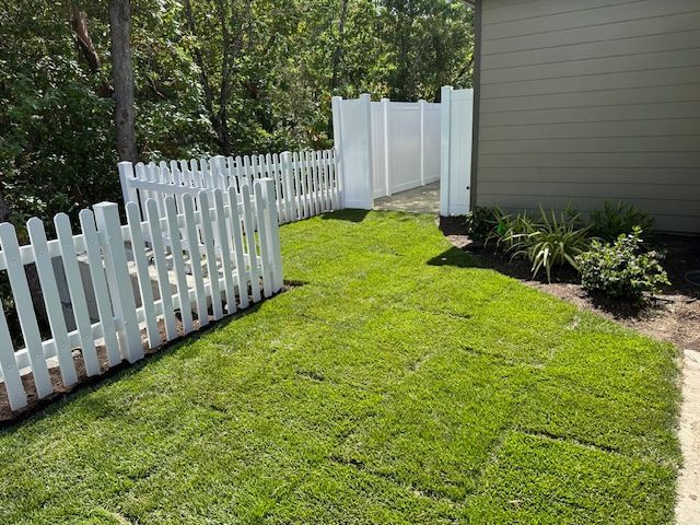 A newly installed patch of green sod lawn beside a house, enclosed by white picket and privacy fences.