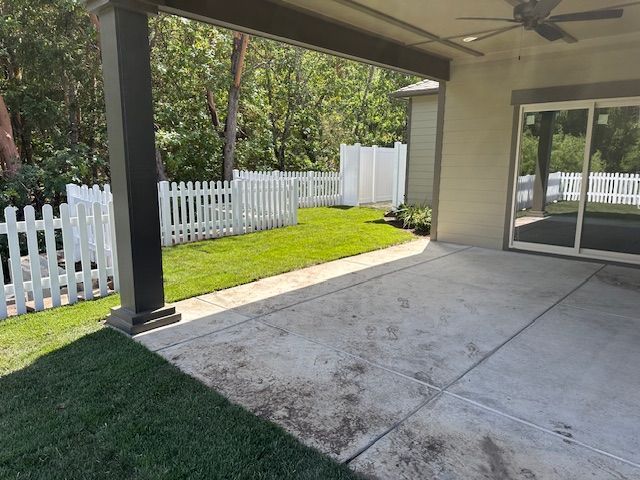 A covered patio featuring a concrete floor, a dark support pillar, and a view of a grassy yard with a white picket fence.