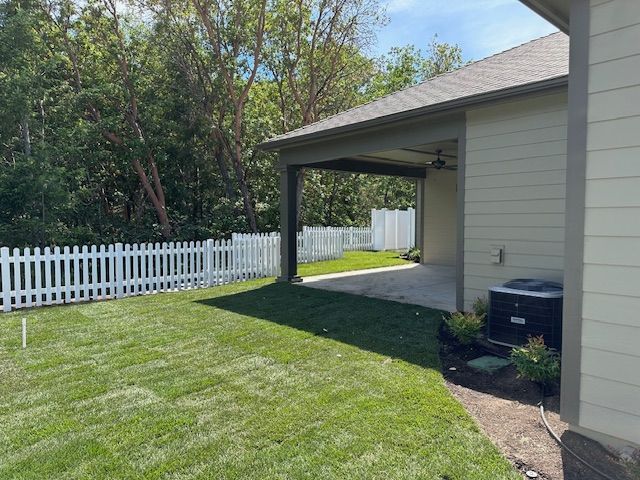 A back patio with a beige exterior, a white picket fence, and a lush green lawn on a sunny day.