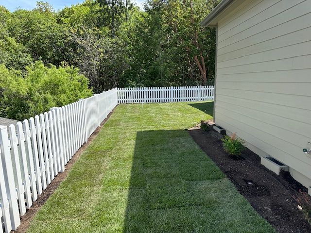 A backyard view featuring a white picket fence, a freshly laid sod lawn, and a side view of a beige house.