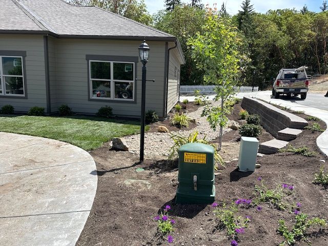 A tan house with a circular concrete driveway, a green utility box, a lamp post, and a stone retaining wall in a yard.