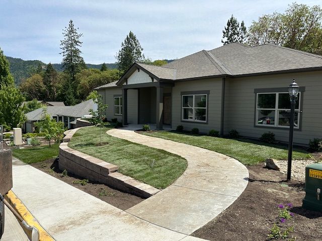 A beige house with a concrete walkway, a stone retaining wall, and a green lawn, set against a backdrop of trees and hills.