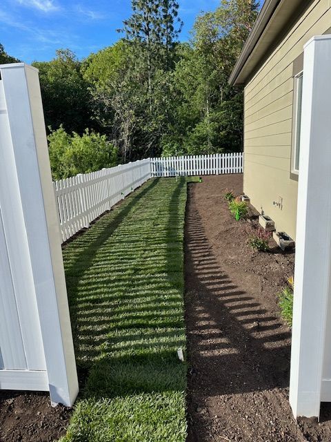A narrow strip of freshly laid sod lawn between a house wall and a white picket fence, with dirt along the house side.