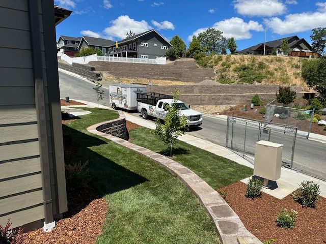 A white pickup truck with a trailer drives up a residential street past a landscaped front yard with a curved stone wall.