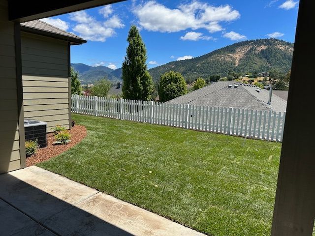 A backyard view from a patio, showing a green lawn, white picket fence, and distant mountains under a blue sky.