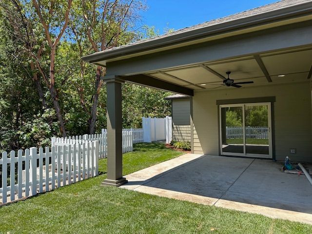 A covered concrete patio with a ceiling fan attaches to a house, featuring a white picket fence and trees in the yard.