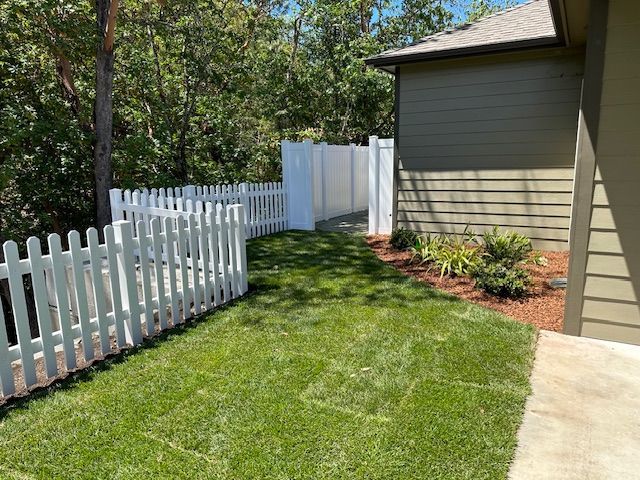 A white picket fence lines a green lawn next to a house with beige siding and a privacy fence leading to trees.