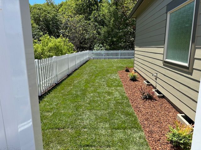 A grassy backyard alongside a tan house, featuring red mulch landscaping, white vinyl fencing, and trees in the background.