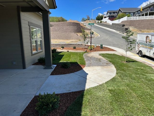 A residential patio with a concrete walkway leads to a landscaped lawn, with a road and a parked truck visible uphill.