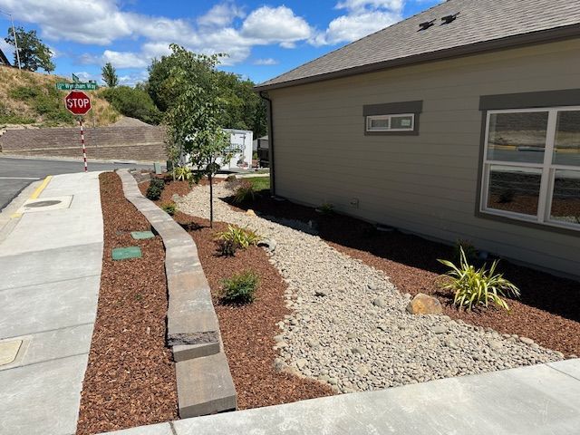 A tan house with a landscaped yard featuring a rock drainage swale, mulch, and a stone retaining wall near a sidewalk.