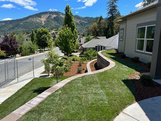 A landscaped residential front yard with a curved retaining wall, a grassy slope, trees, and a mountain view in the distance.