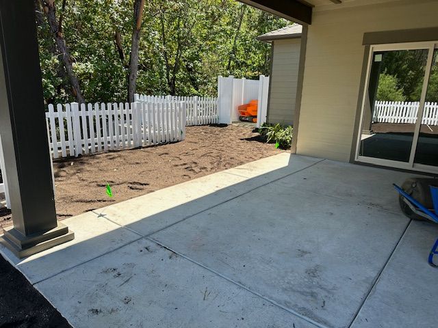 A concrete patio looks out onto a yard with bare dirt, a white picket fence, and a distant orange wheelbarrow.