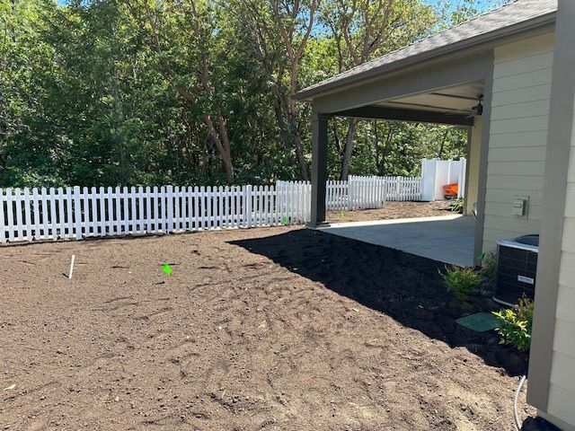 A view of a backyard with fresh dirt, a white picket fence, and a covered concrete patio attached to a house.