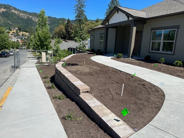 A concrete retaining wall borders a curved walkway leading to a house, with fresh soil and green landscaping flags.