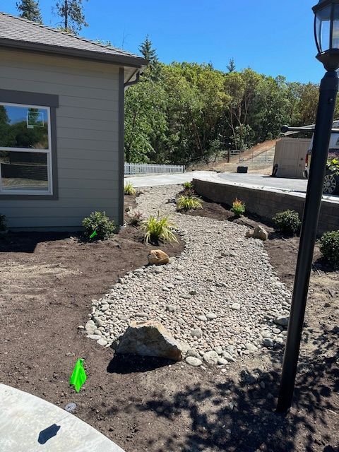 A stone-lined drainage swale curves through a landscaped yard next to a tan building on a sunny day.