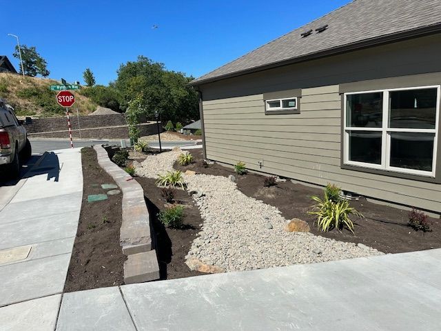 A dry creek bed of light-colored gravel runs along the side of a tan house, separated from a sidewalk by a low stone wall.