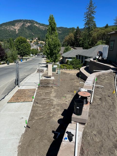A construction site along a sidewalk and retaining wall with exposed dirt, utility markings, and residential houses.