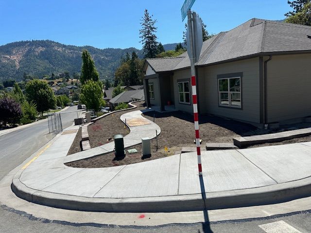 A tan house with a sidewalk winding through a mulched front yard at a street corner with mountains in the background.