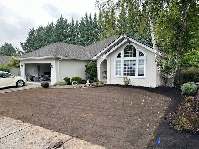Single-story beige house with a grey roof and large front window, featuring a freshly tilled dirt lawn.