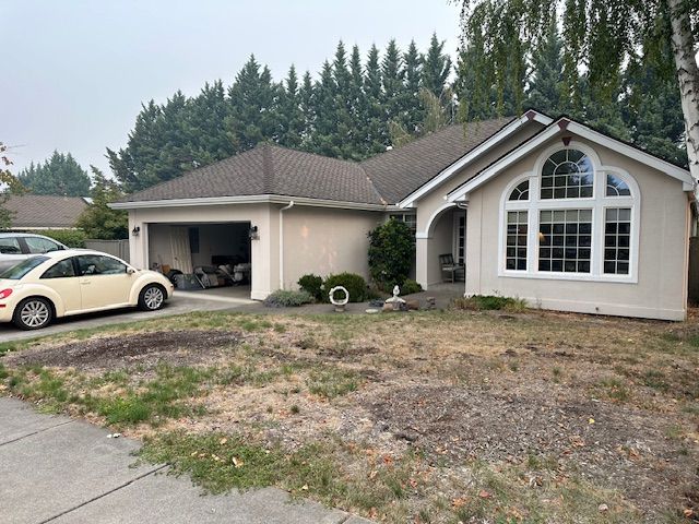 A single-story tan house with a two-car garage, a large arched window, and a dry, patchy front lawn.