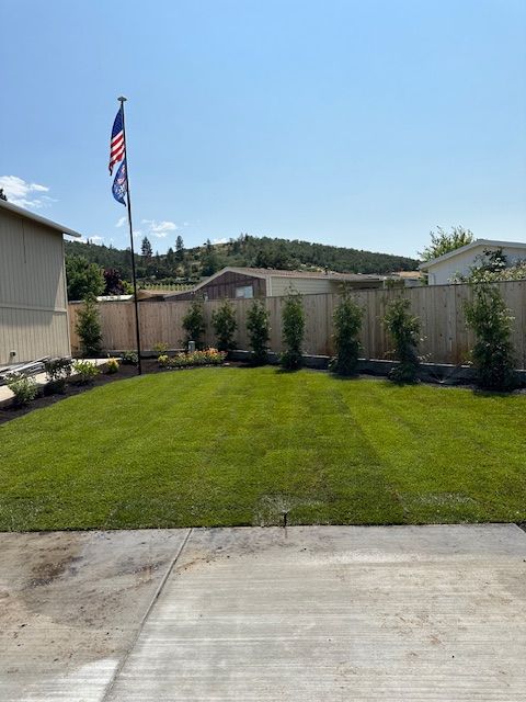 A manicured green lawn framed by small evergreen trees along a wooden fence, with an American flag flying on a pole.
