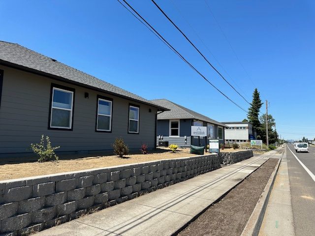 Gray houses with dark trim sit behind a low stone retaining wall along a sidewalk and a paved road on a clear sunny day.