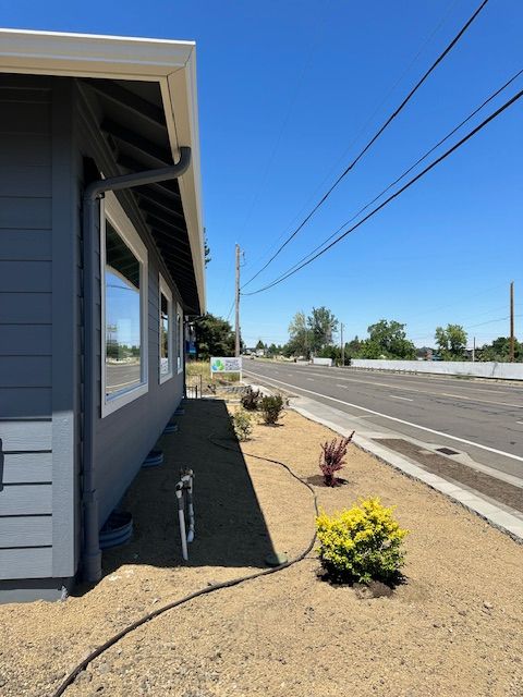Gray building exterior with a downspout and small shrubs planted in gravel alongside a road under a bright blue sky.