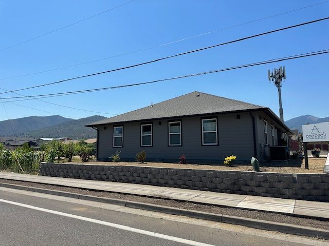 Gray, single-story commercial building with a dark roof behind a block retaining wall, set against a backdrop of mountains.