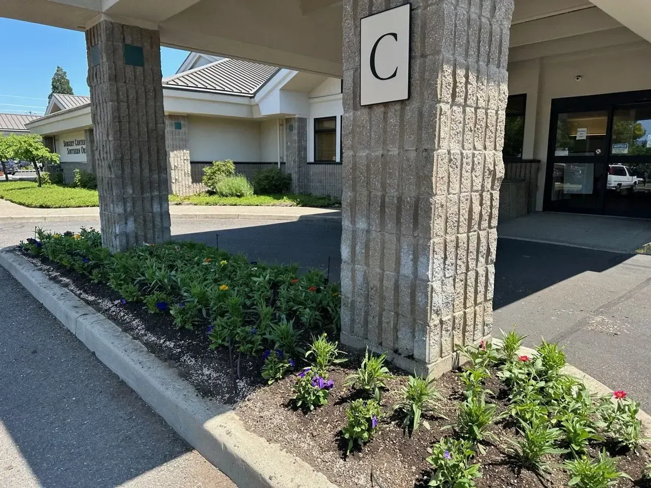 Entrance C of a building featuring brick support columns and a garden bed with small flowers in the foreground.