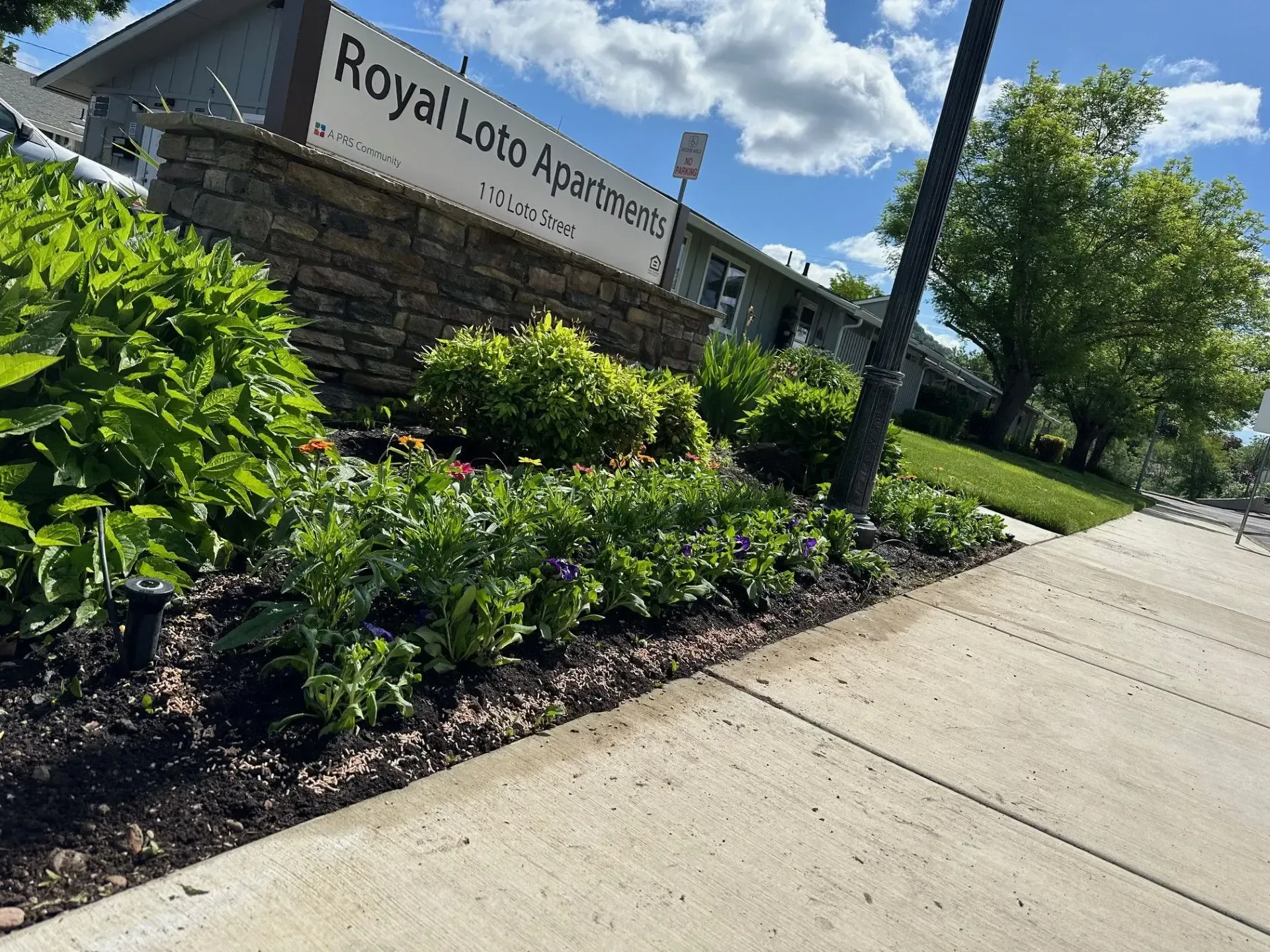 The Royal Loto Apartments sign and a stone wall behind a landscaped flower bed next to a sidewalk on a sunny day.