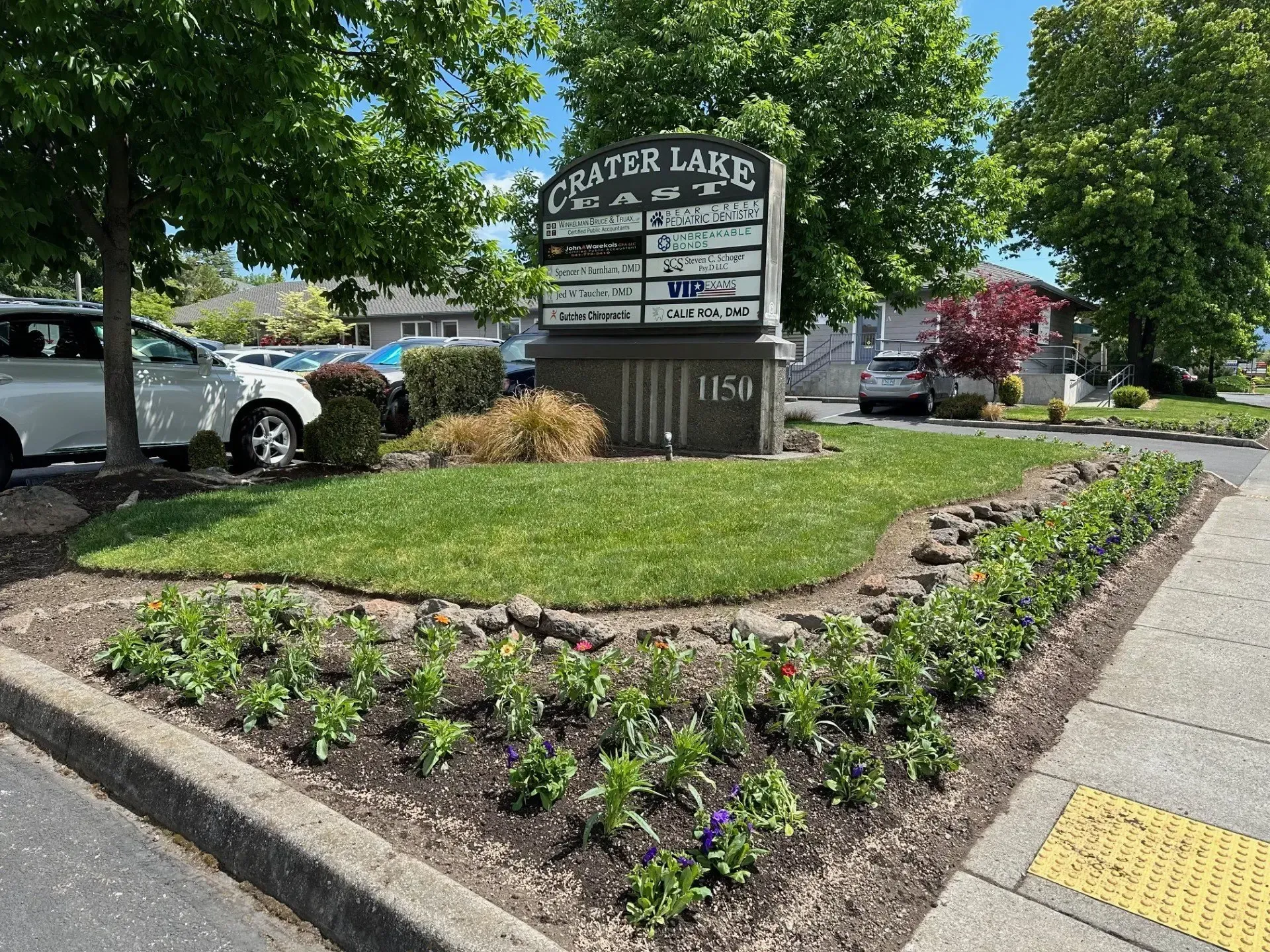 A stone sign for Crater Lake, surrounded by a lawn and a garden bed of small flowers, near a street with parked cars.
