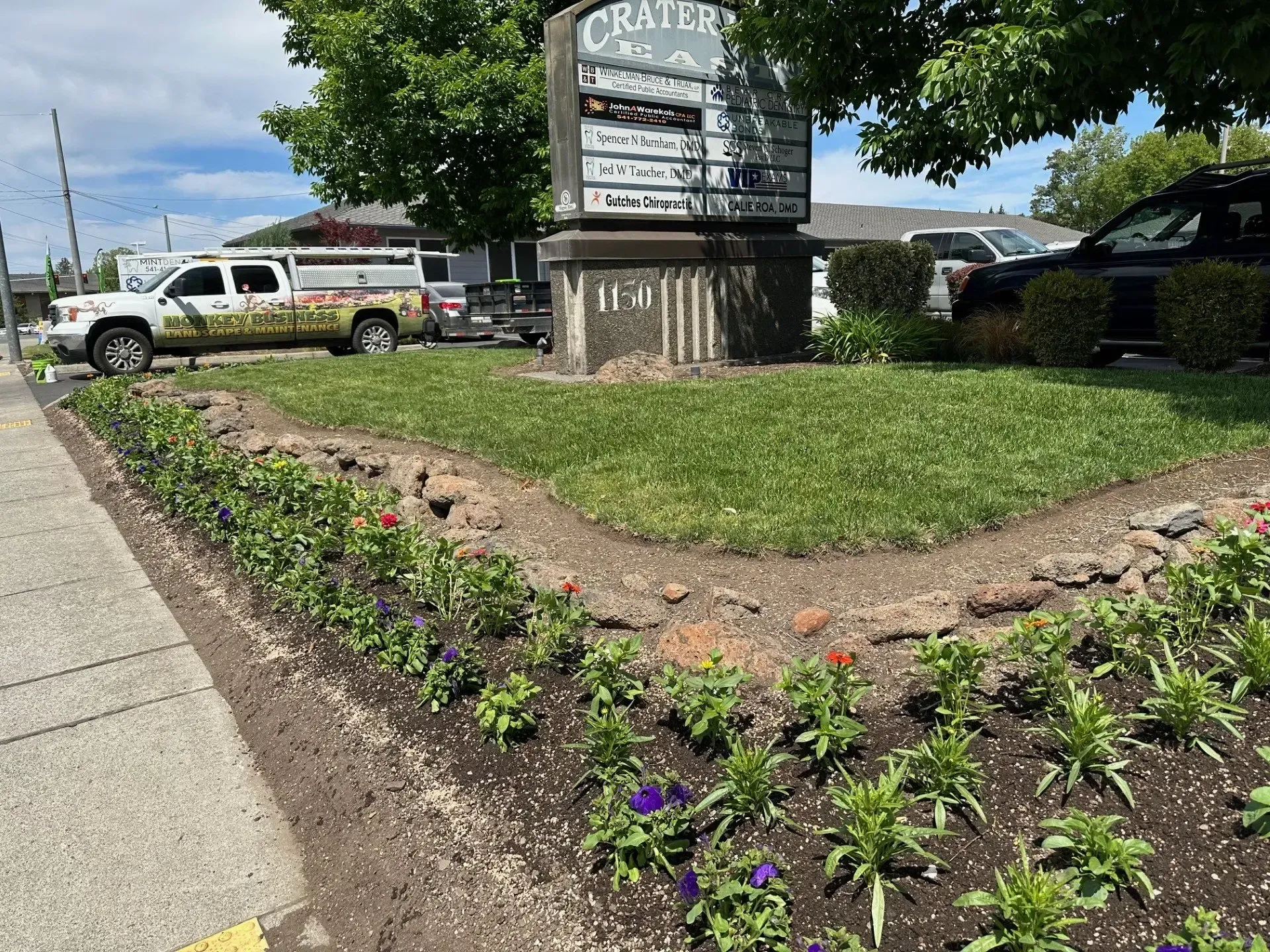 A sign for Crater Lake, flanked by a lawn and a sidewalk-side flower bed with small blooming flowers, next to a truck.