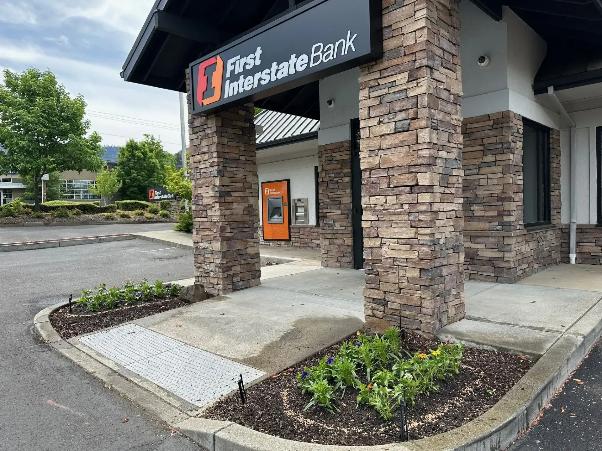 The First Interstate Bank entrance featuring stone columns, an orange ATM, and landscaping beds on a sunny day.
