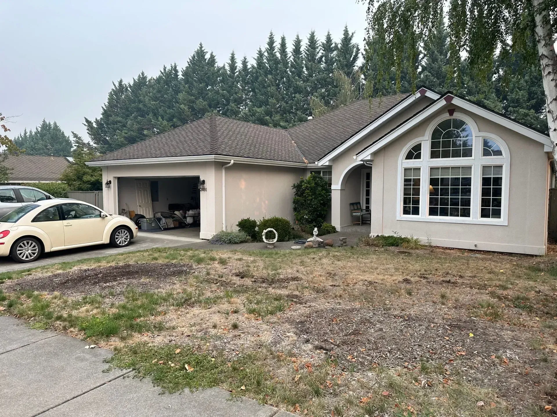 A light beige single-story house with a two-car garage and a large arched window, viewed from a dry, patchy front lawn.