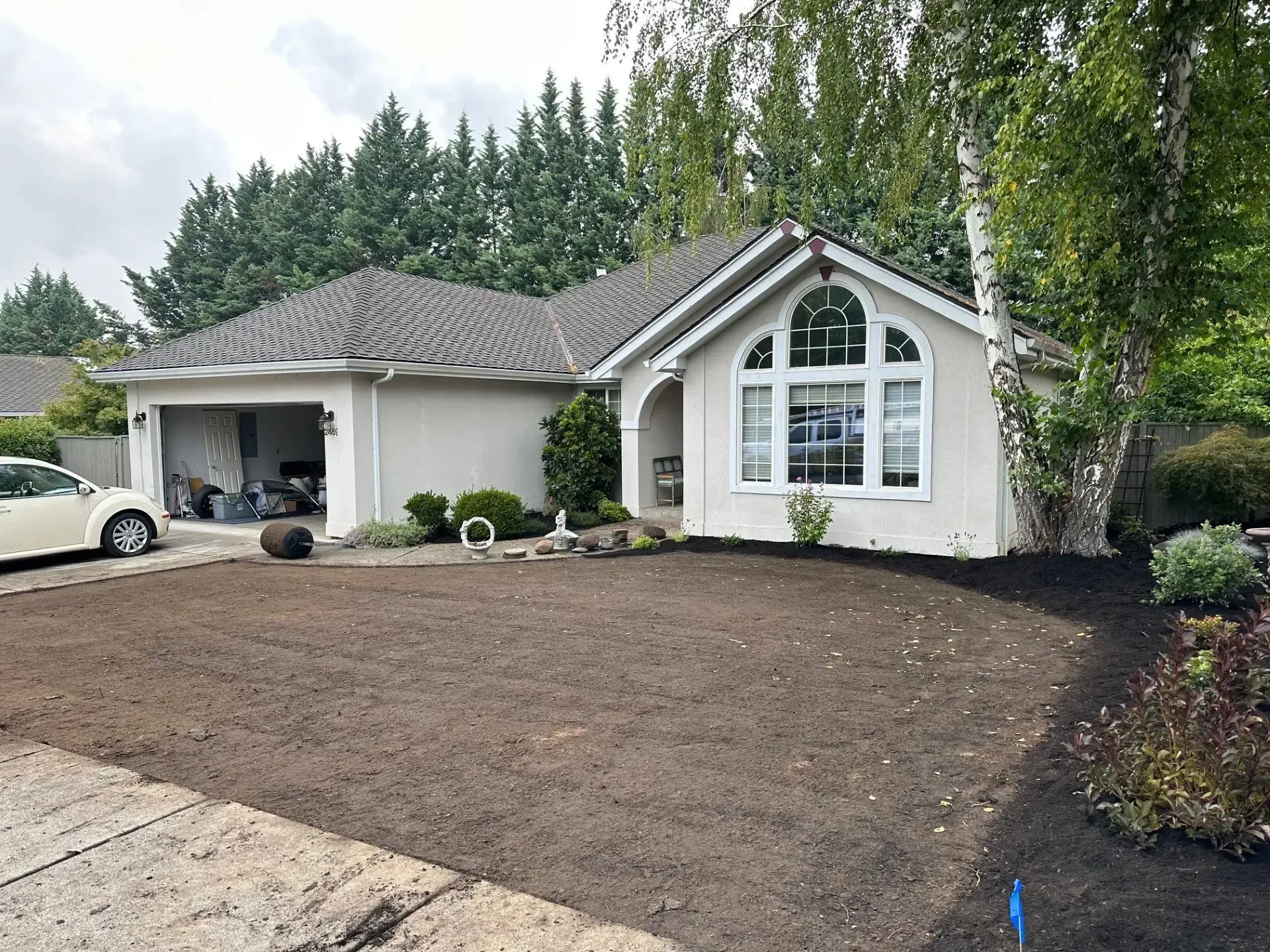 A suburban, single-story light-colored house with a garage and a newly prepared dirt lawn in the front yard.