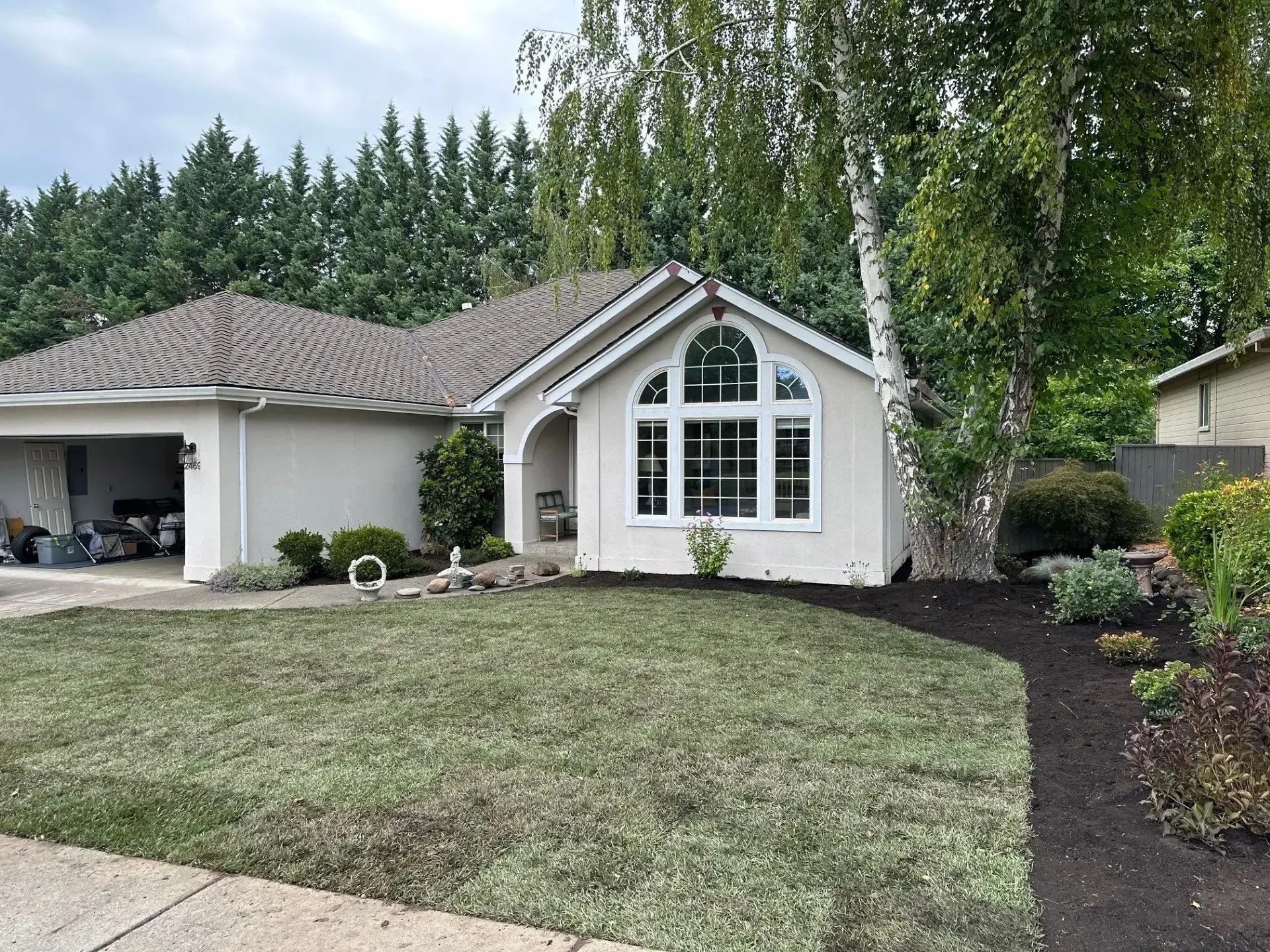A single-story, beige suburban home with a two-car garage, a large front window, and a fresh mulch bed in the front yard.