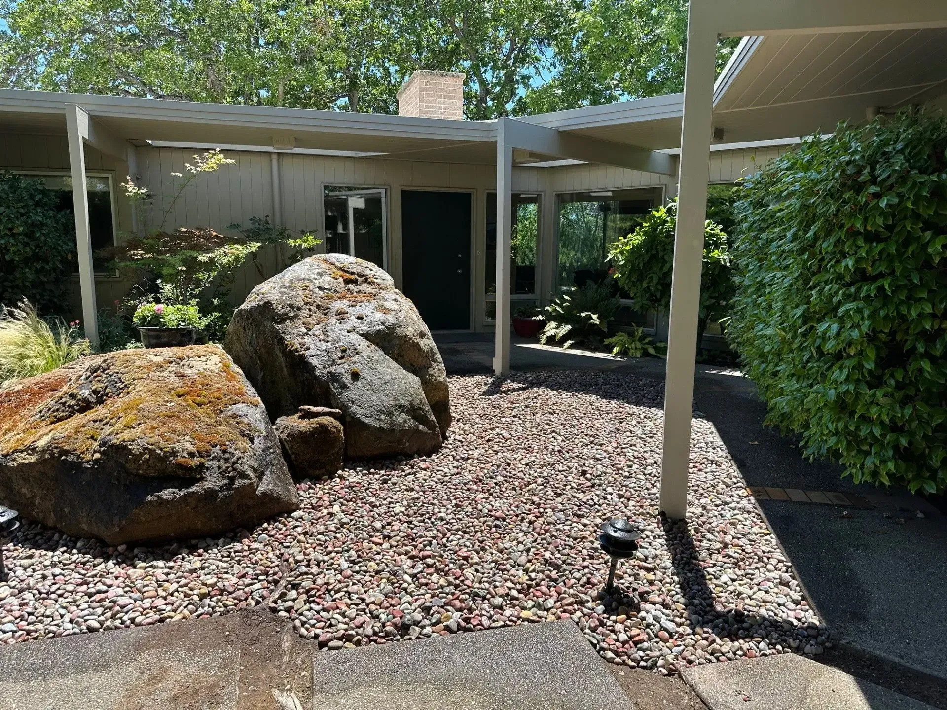 A modern home entrance with a pebble courtyard featuring two large boulders under a covered walkway and greenery.