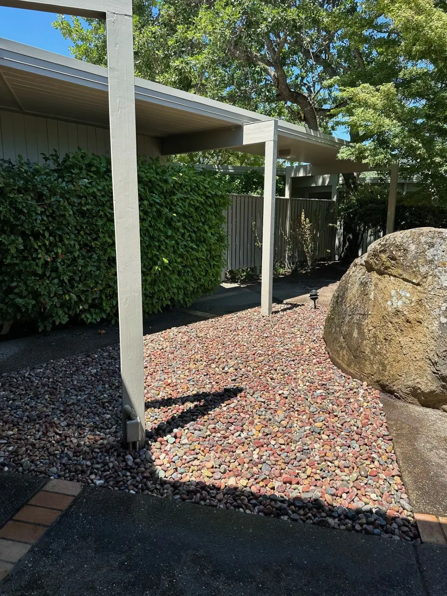 A walkway covered in multicolored gravel leads past a large rock and a hedge toward a covered patio area under trees.