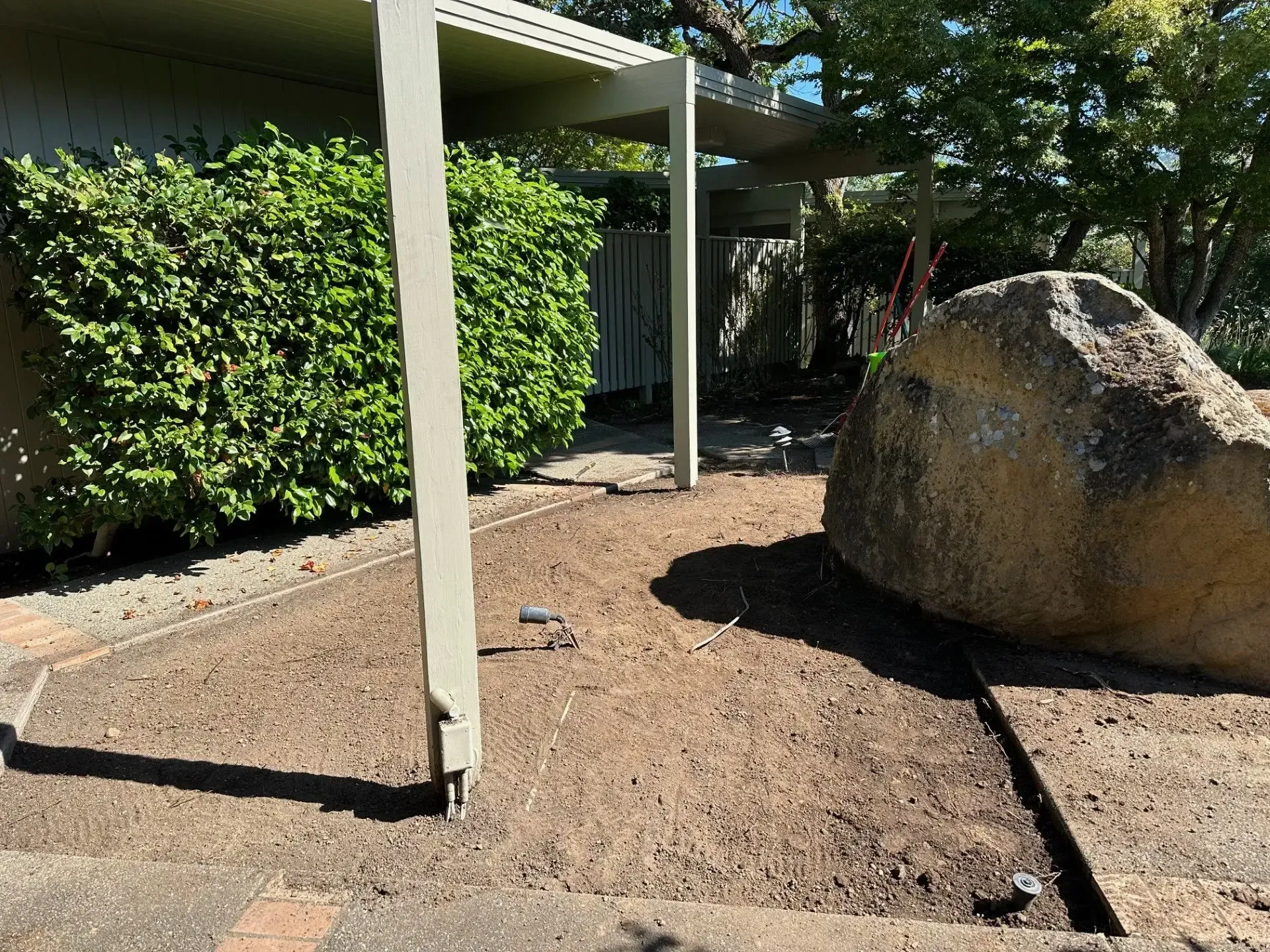A patch of brown, bare garden soil next to a large tan boulder and a light-colored patio post, bordered by a green hedge.