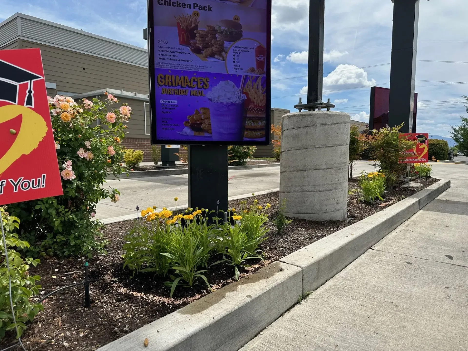 A digital drive-thru menu board standing in a landscaped island with yellow flowers and a concrete bollard.
