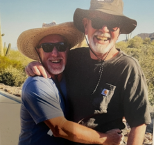 Two smiling men in hats and sunglasses embrace outdoors in a sunny desert landscape.