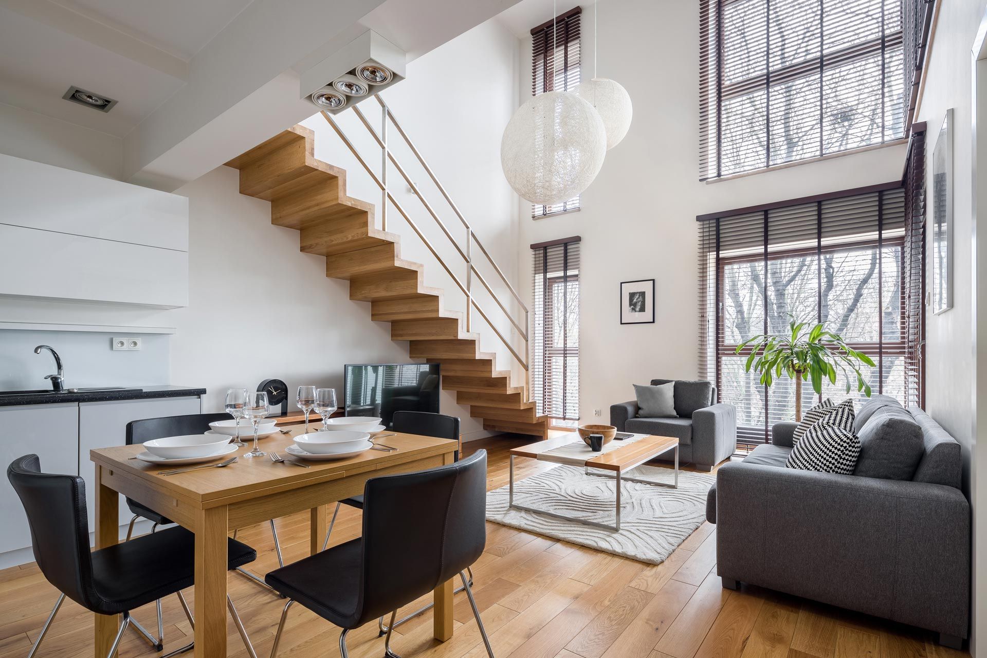 Modern open-plan living room with wooden staircase, dining table, gray sofa, and large windows