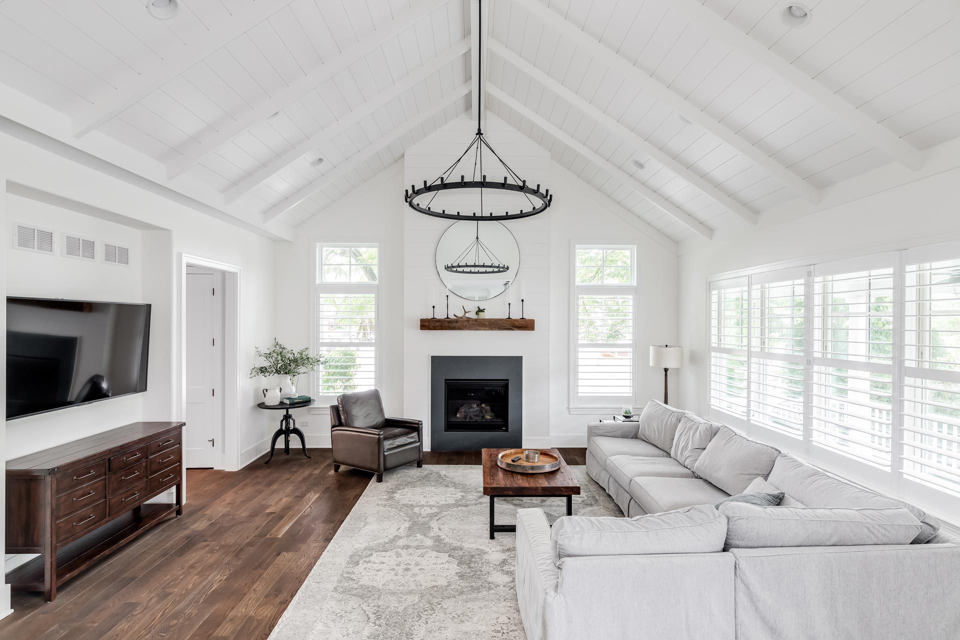 Bright white living room with vaulted ceiling, fireplace, gray sofa, and dark wood floors.