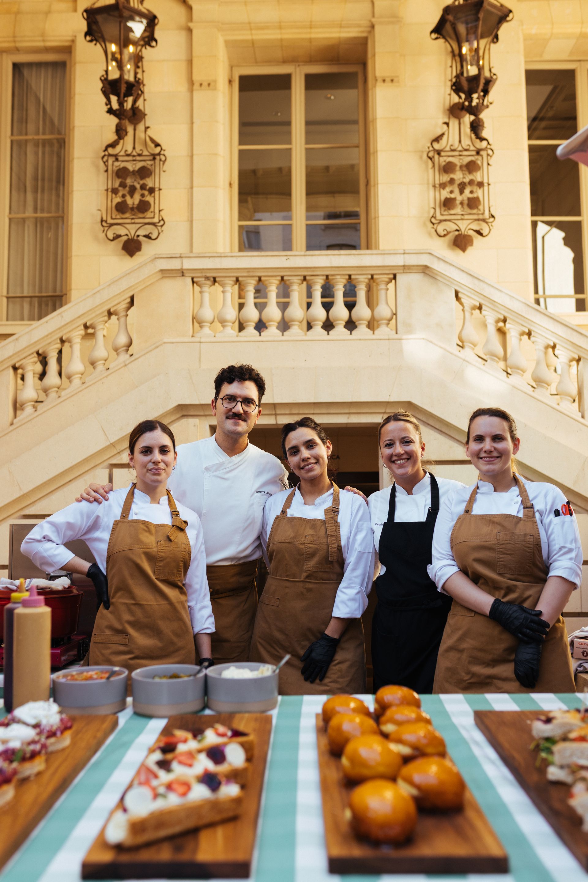 Un equipo culinario profesional, ataviado con batas blancas y delantales marrones, posa frente a una gran y ornamentada escalera con comida gourmet.