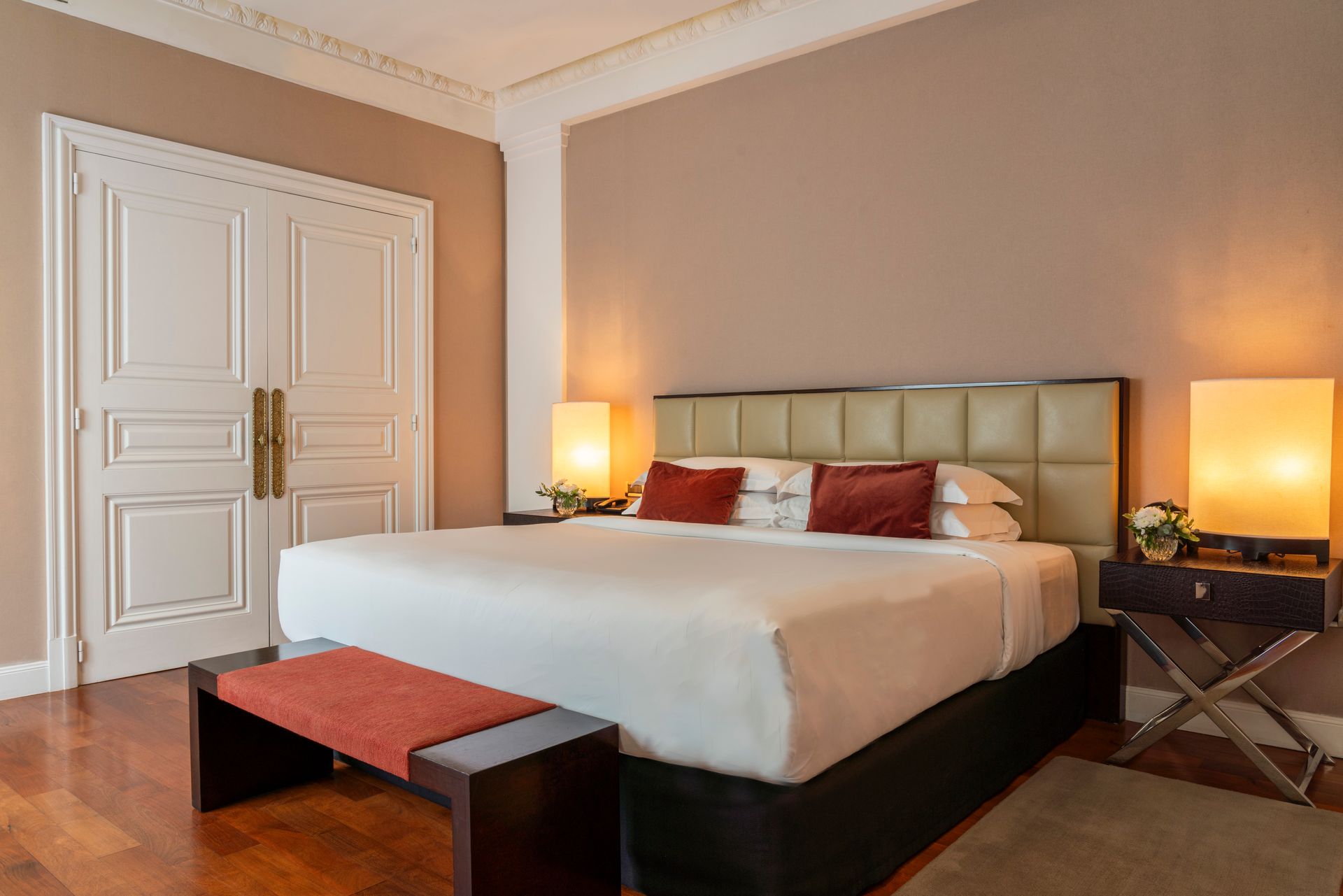 Una habitación de hotel con una cama grande, dos lámparas de noche y un banco, con paredes en tonos neutros y suelo de madera.