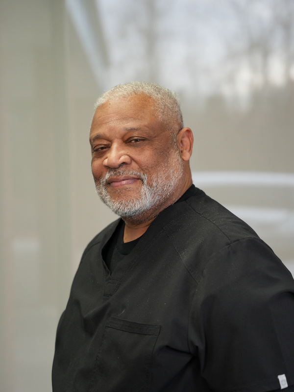 An older Black man with a gray beard smiles, wearing a black uniform, in a bright room.