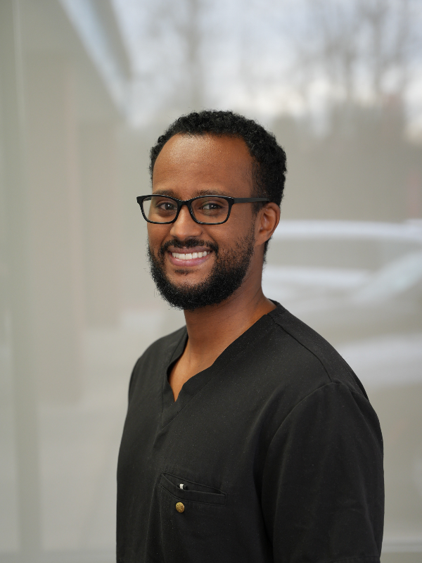 A smiling man with a beard and glasses wearing a black scrub top, looking at the camera.