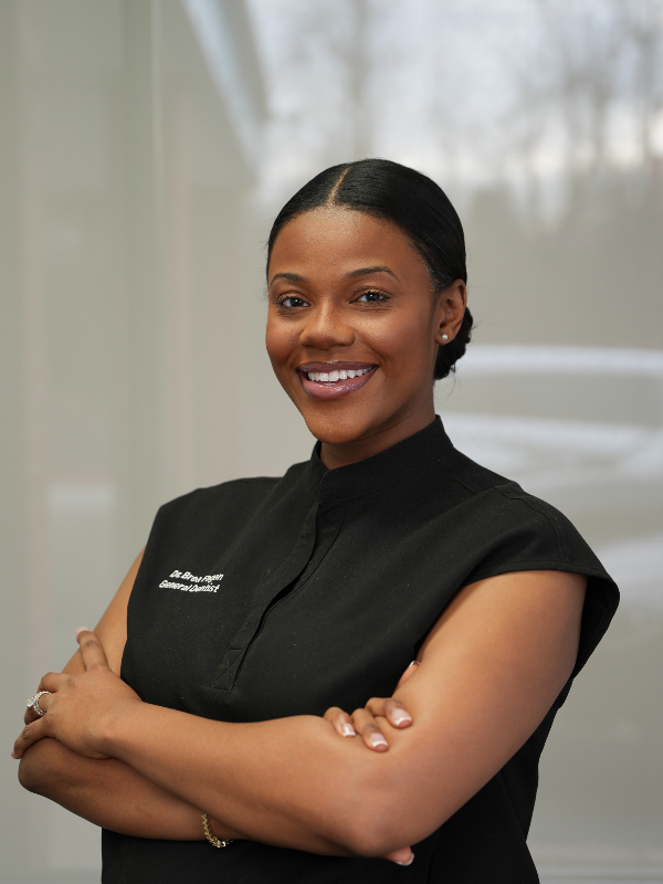 A Black woman in a black uniform smiles with crossed arms, indoors.